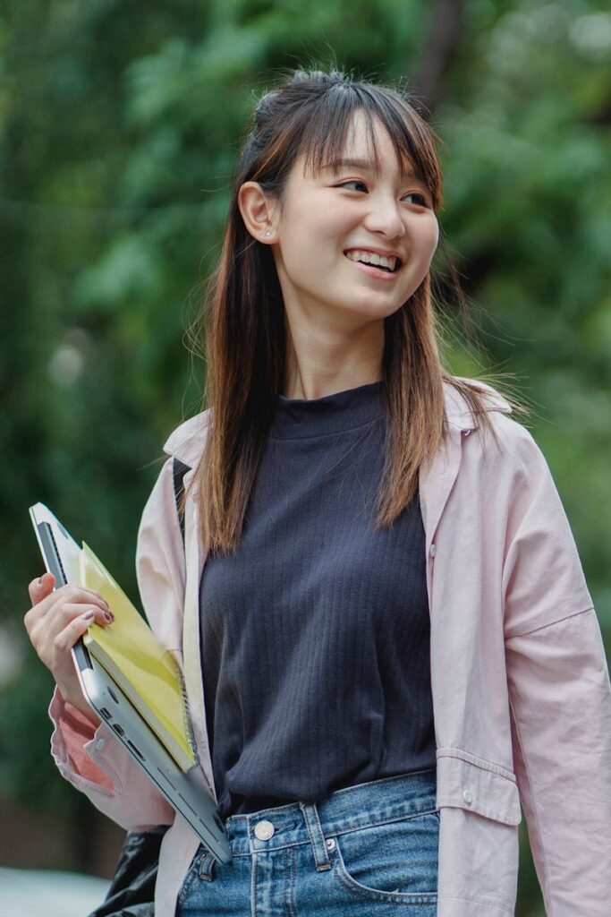pexels-photo-5538001-5538001 Smiling young Asian woman carrying a laptop and books, enjoying a sunny day outdoors.