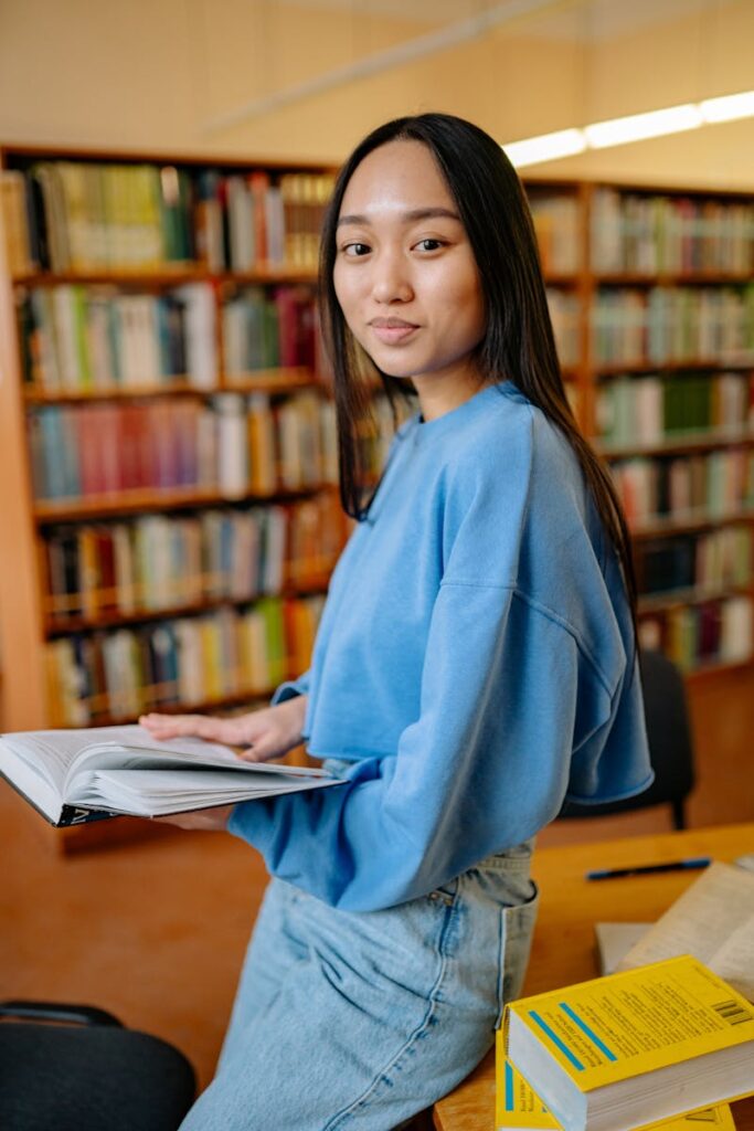Asian woman reading a book in a library, smiling while studying surrounded by shelves.